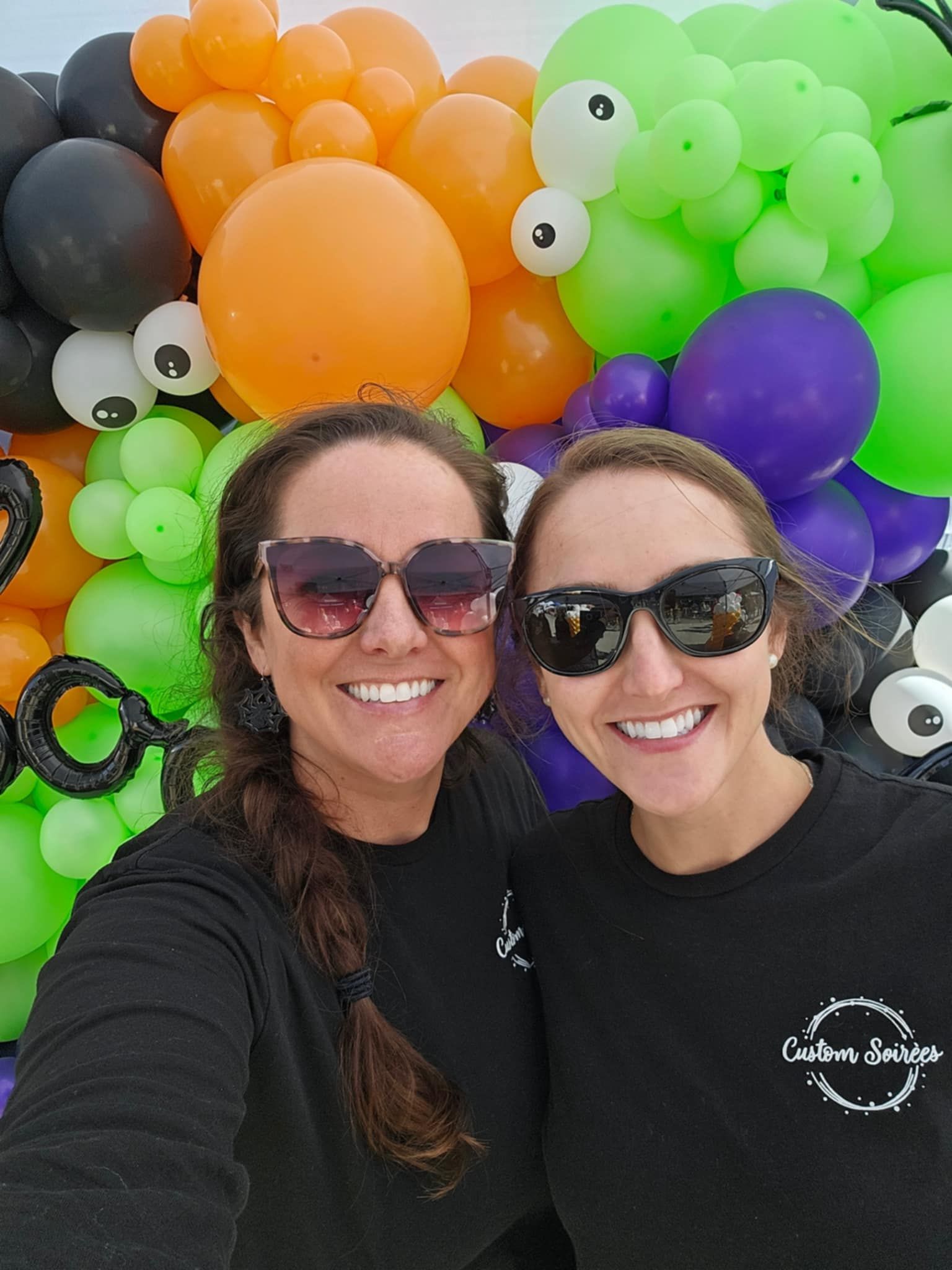 Two women wearing sunglasses are posing for a selfie in front of a wall of balloons.