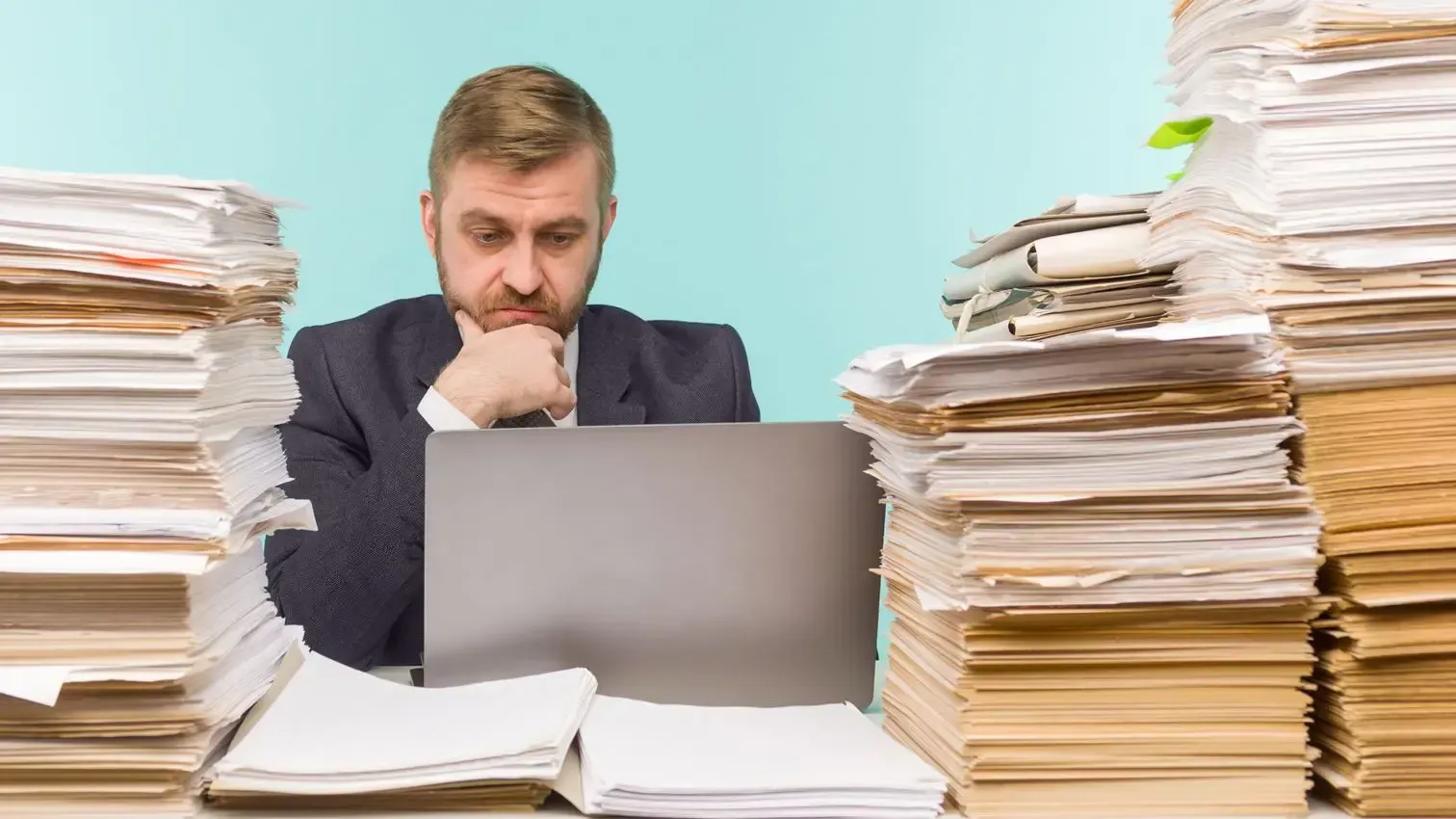 Uma pessoa de terno está sentada em uma mesa com grandes pilhas de papel de cada lado, olhando pensativamente para a tela de um laptop.