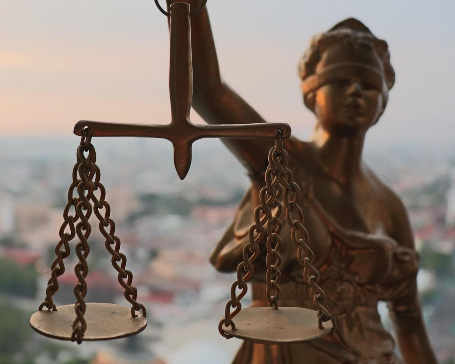 Uma estátua de bronze da Dama da Justiça segurando uma balança, com uma paisagem urbana desfocada ao fundo.
