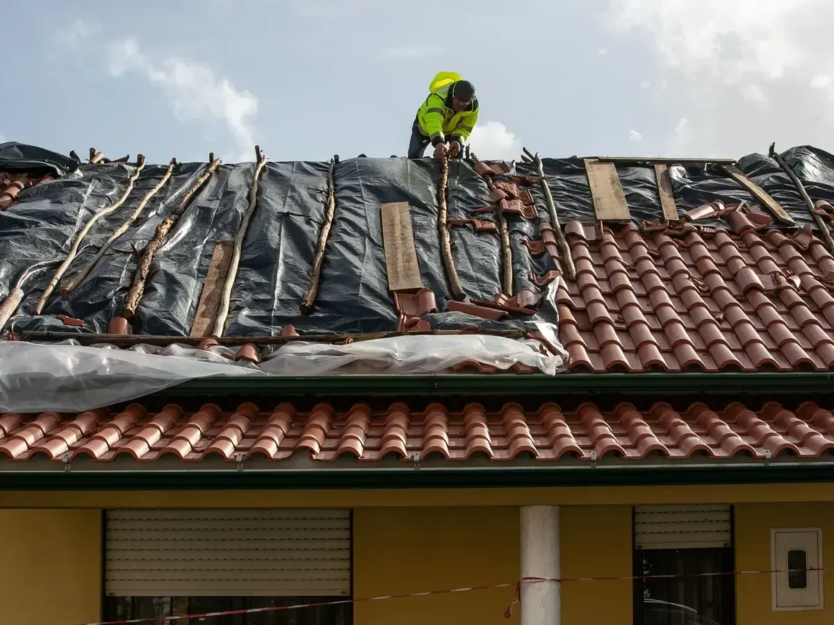 Uma pessoa vestindo uma jaqueta amarela brilhante trabalha em um telhado, instalando uma membrana protetora preta sobre ripas de madeira.