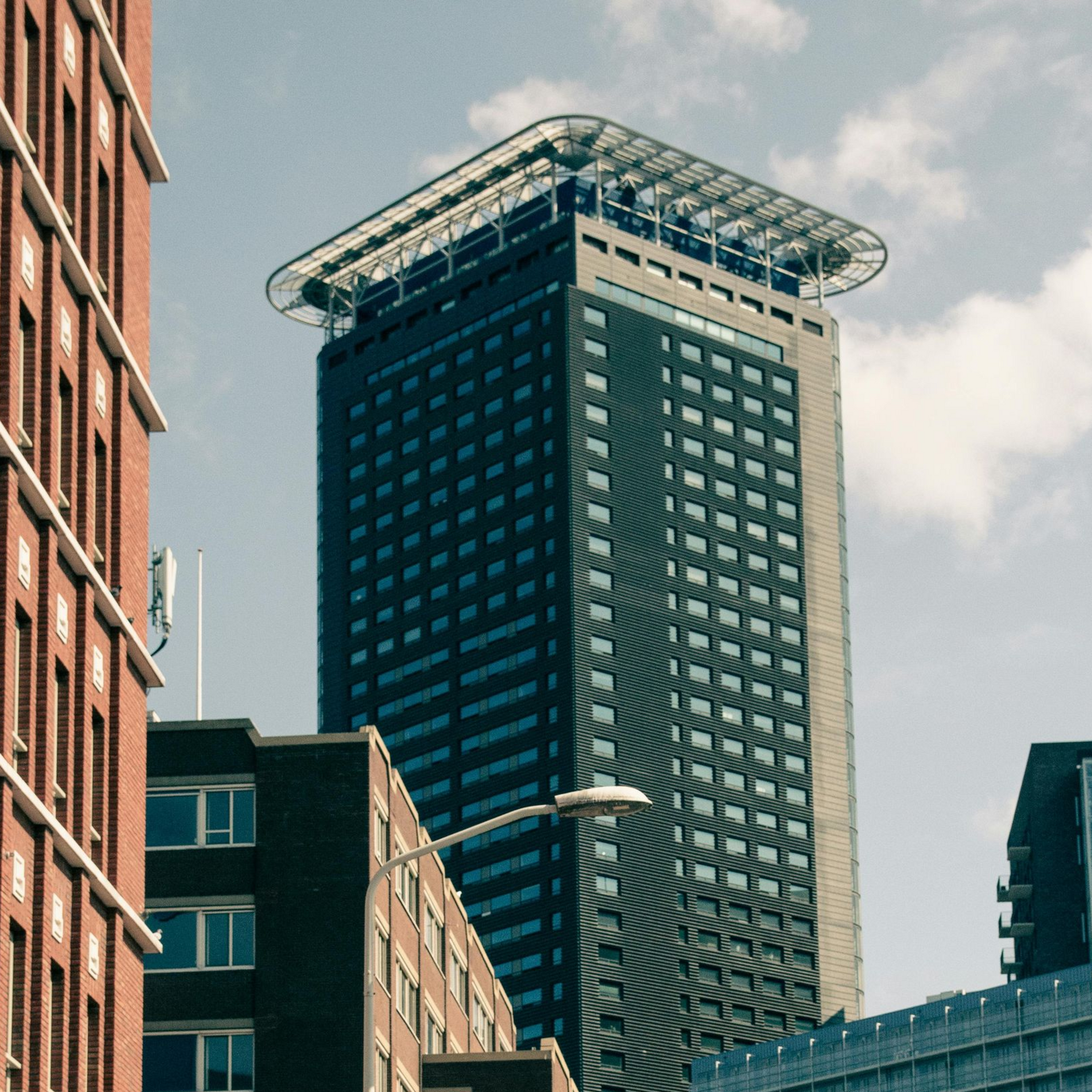 Um arranha-céu moderno, com fachada escura e uma estrutura de cobertura distinta e saliente, ergue-se em meio aos edifícios urbanos sob a luz do dia.