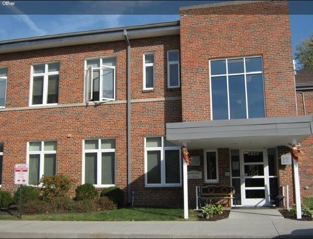 Two-story brick building with white-framed windows, gray canopy over entrance, and small garden in front.