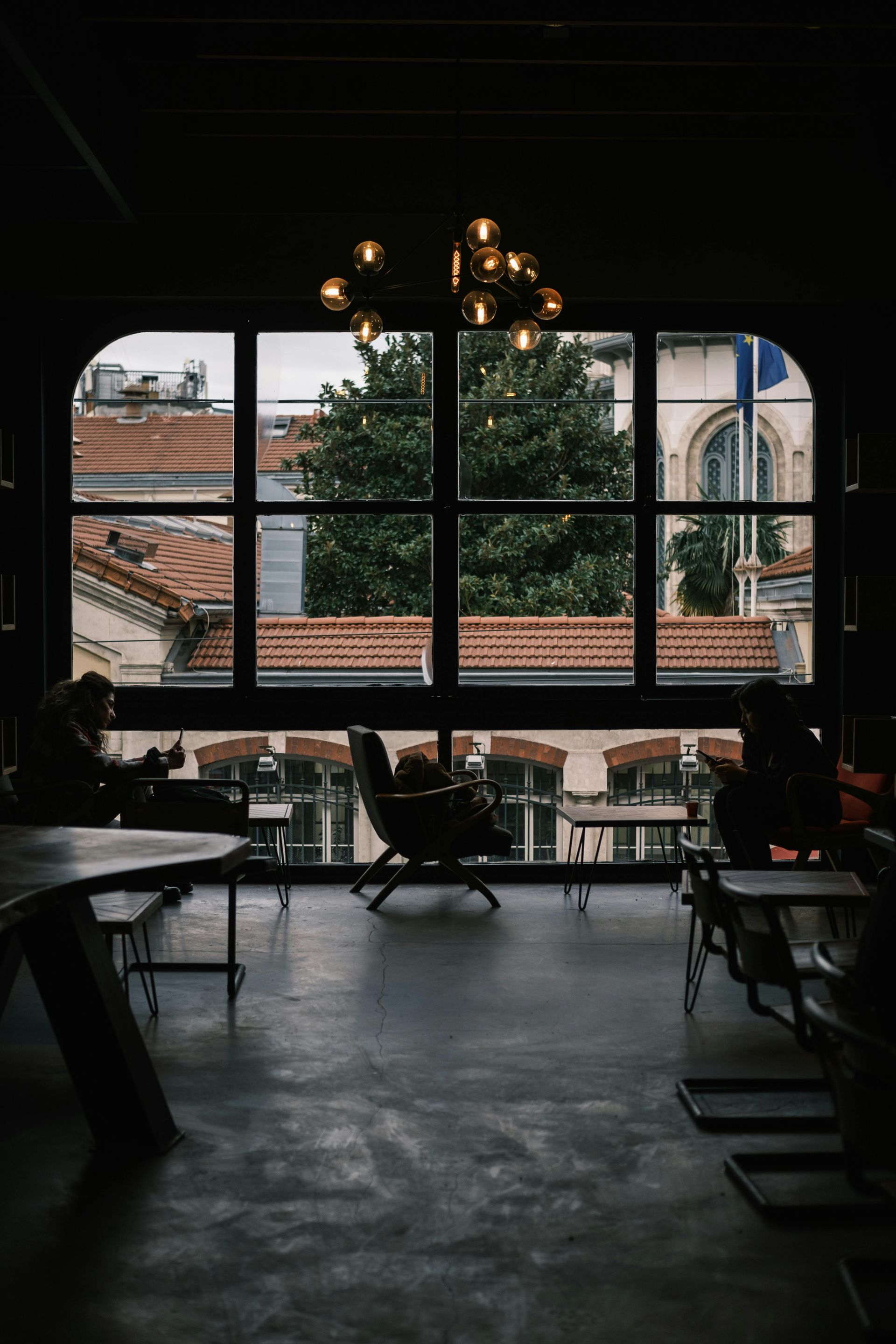 Interior of a room with a large window overlooking buildings. People sit at tables, dimly lit.
