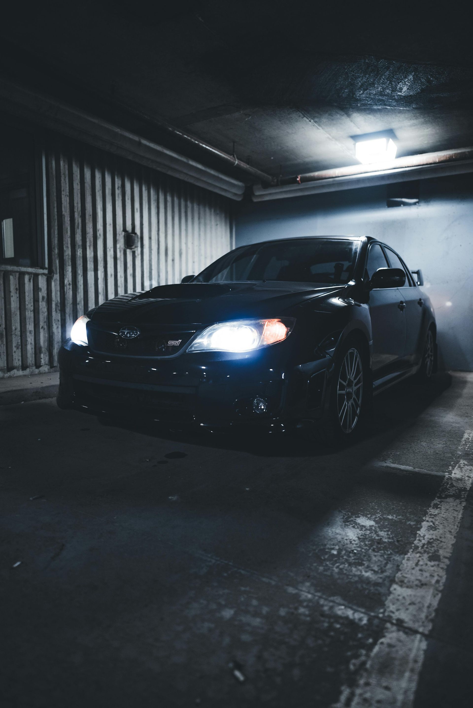 Black Subaru car parked in a dark garage, headlights on, with a blurred background.