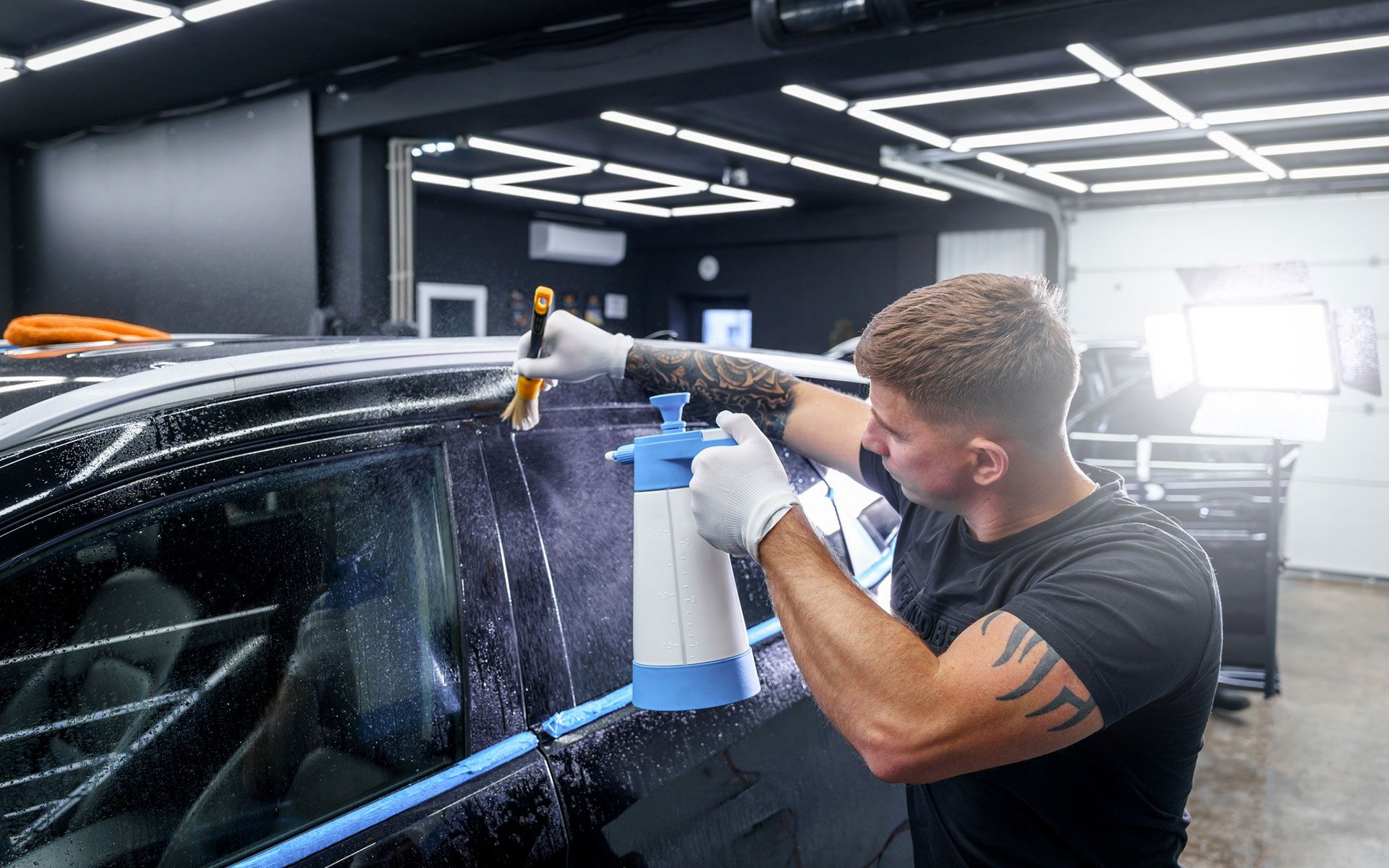 A man is washing a car with a spray bottle in a garage.