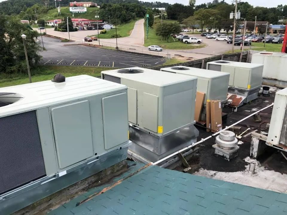 Row of large rooftop HVAC units and vents on a building roof near a roadway, with trees and parking lot beyond