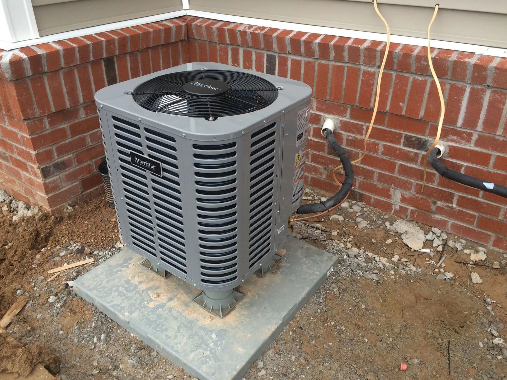 Outdoor HVAC unit beside a brick wall on a concrete pad, with dirt ground and exposed pipes.