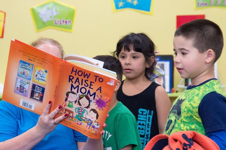 A woman is reading a book to a group of children titled how to raise a mom