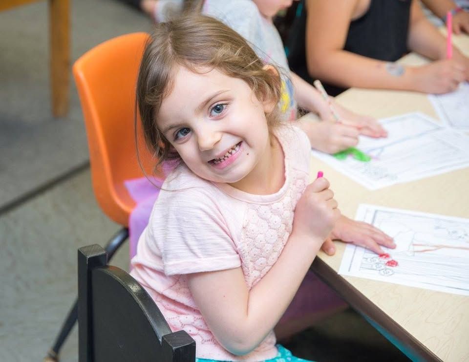 A little girl is sitting at a table in a classroom with other children.