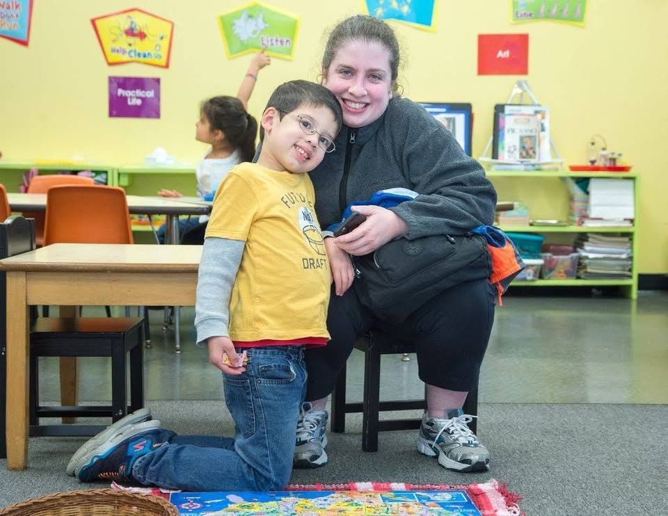A woman sits on a stool next to a young boy in a classroom