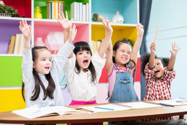 A group of children are sitting at a table with their hands in the air.