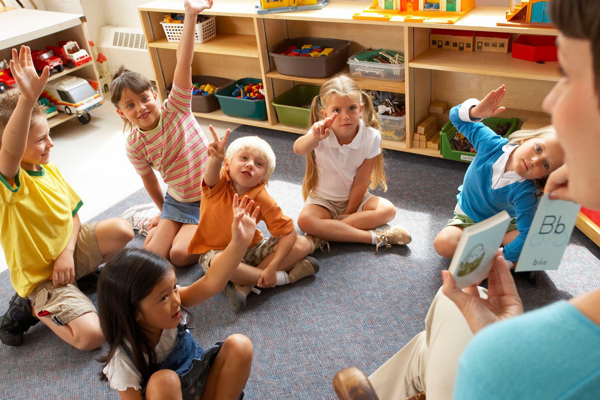 A group of children are sitting on the floor while a teacher holds up a card that says bb