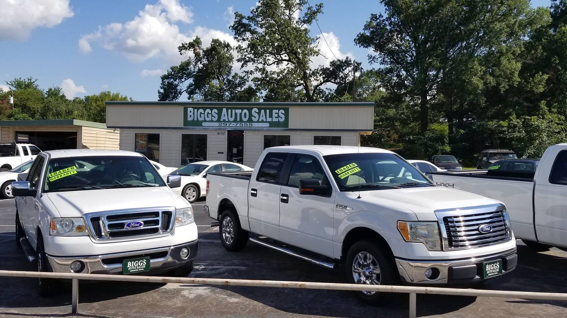 White pickup trucks parked at Bigg Auto Sales, a car dealership, under a blue sky.