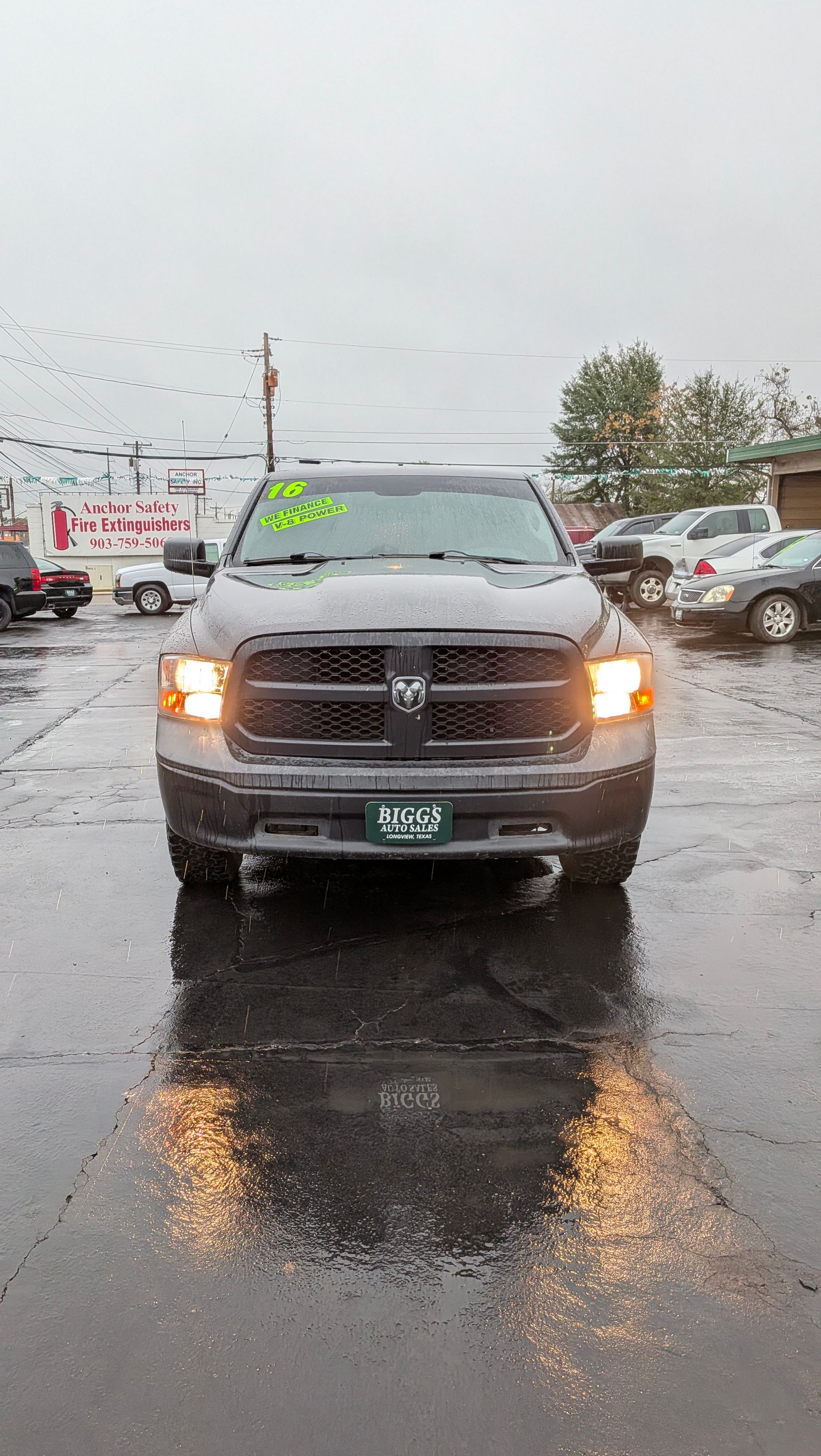 Gray Dodge Ram truck parked on wet pavement, headlights on, cloudy day.
