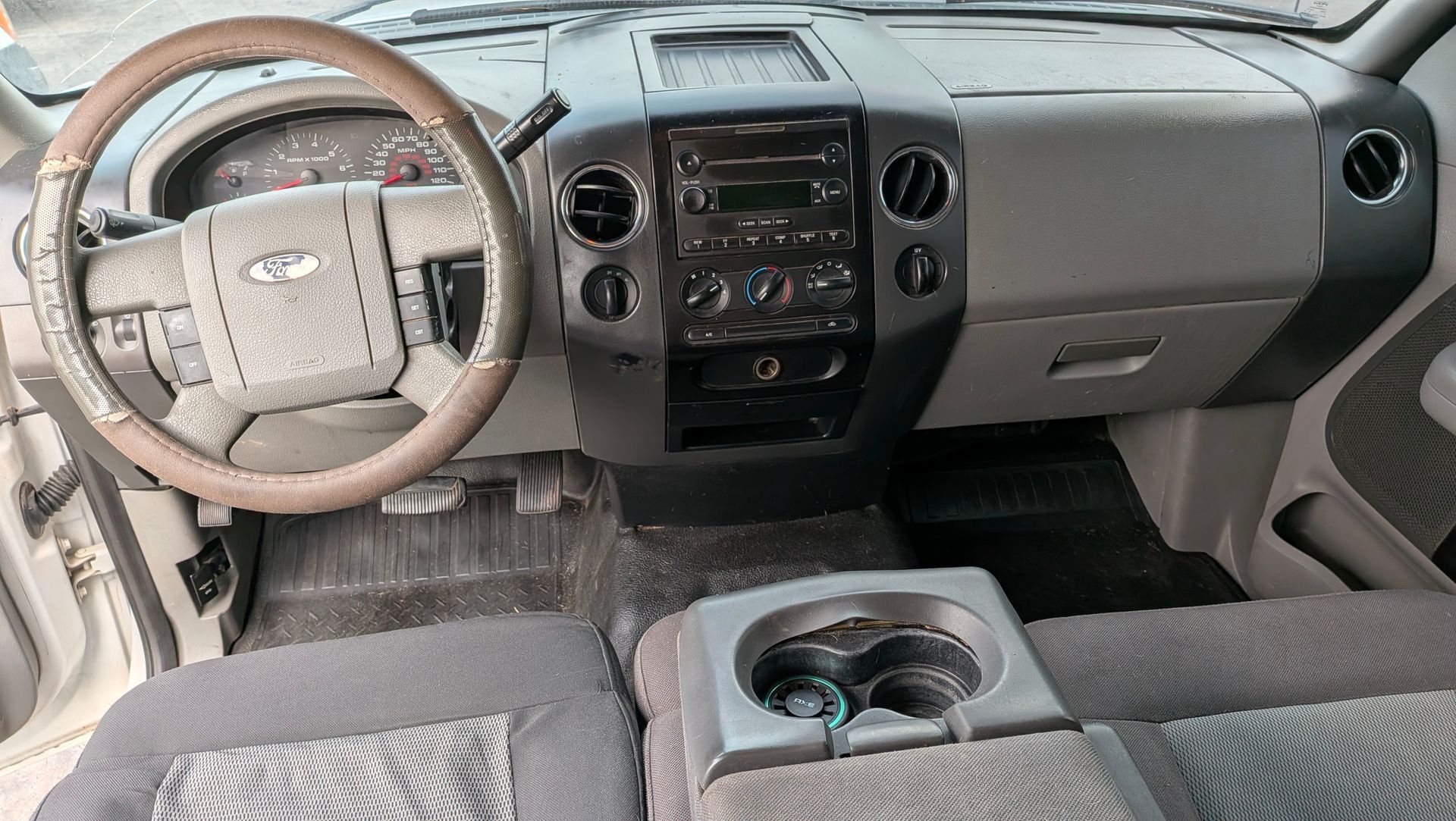 Interior of a truck: dashboard, steering wheel, seats, and cup holders visible. Gray and black color scheme.