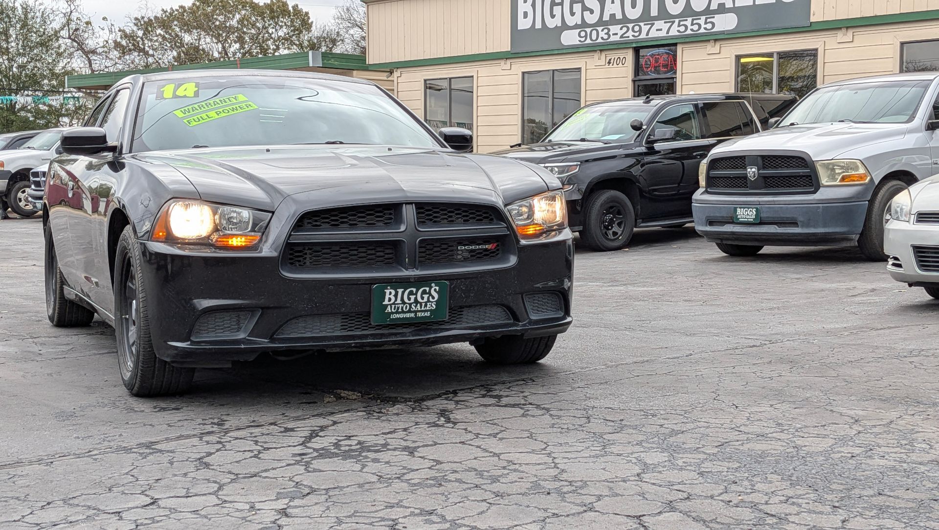 Black Dodge Charger in front of a car dealership, headlights on, wet pavement.