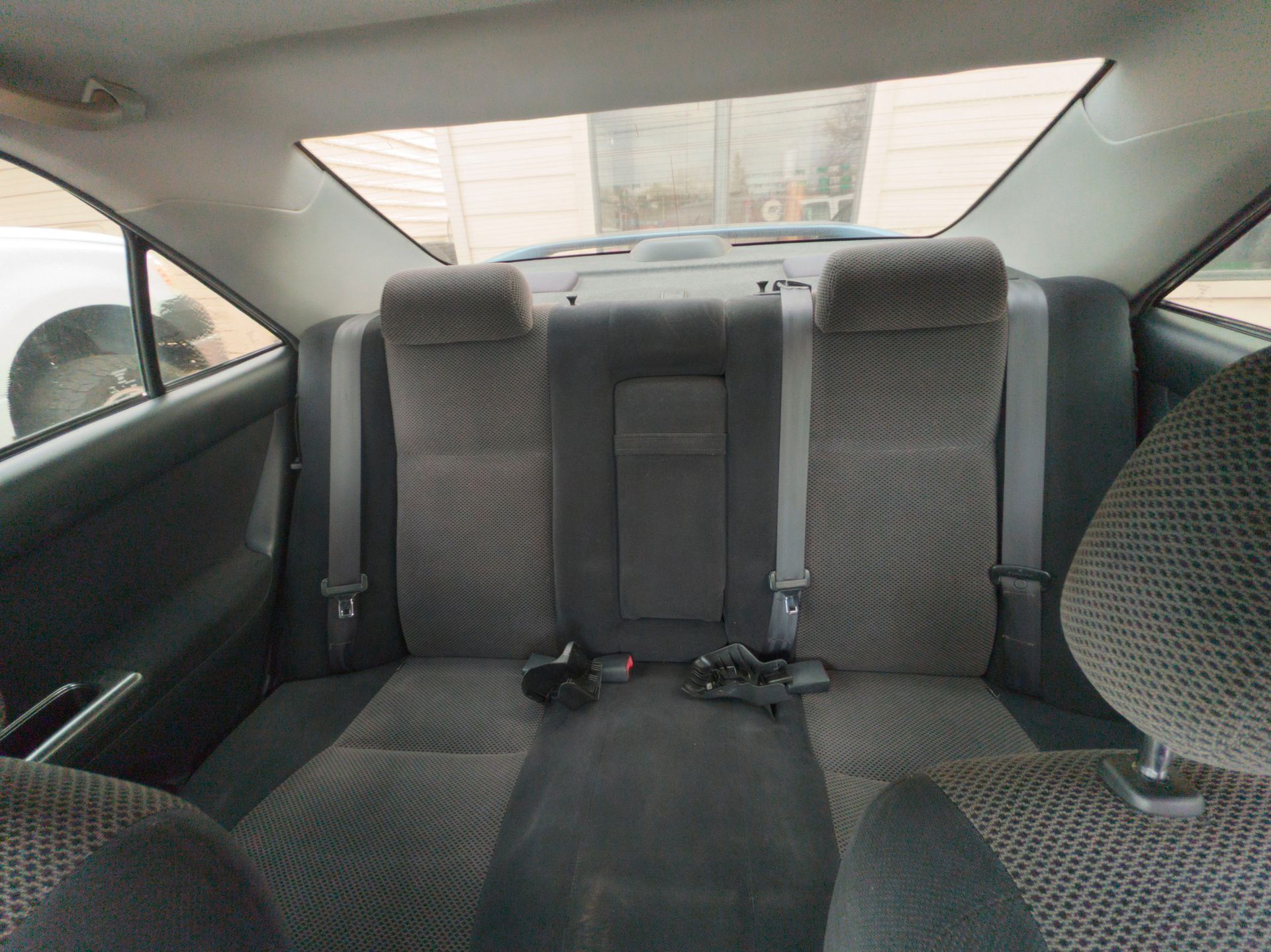 Rear interior of a car, showing the back seat. Gray patterned upholstery, seatbelts, and a window.