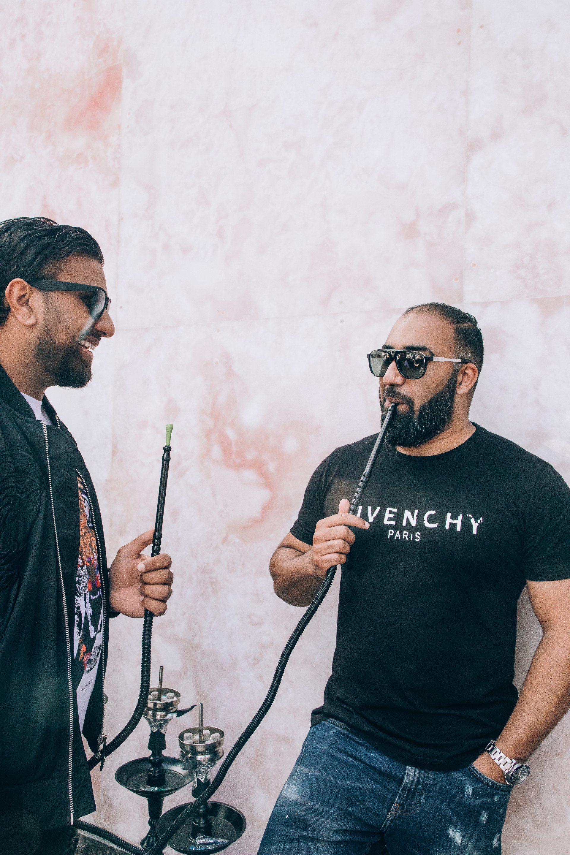 Two men in sunglasses smoking hookah near a textured pink wall. One wears a Givenchy shirt.