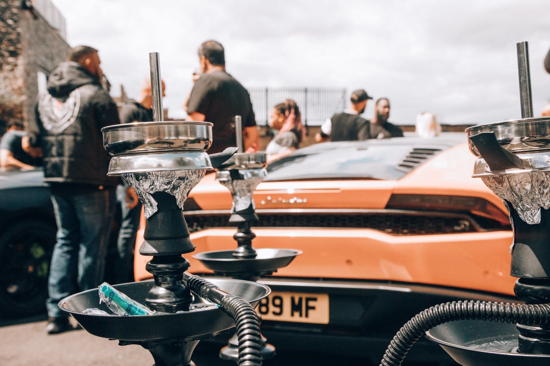 Hookahs in foreground with a blurred orange Lamborghini and people in the background on a sunny day.