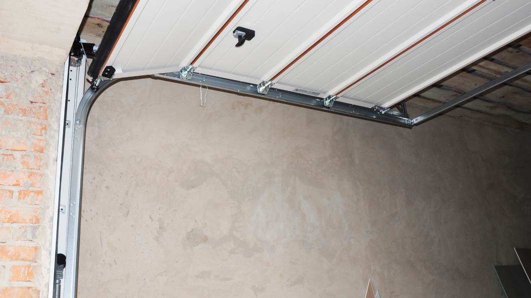 A close-up view of a white, paneled garage door mechanism showing the metal track and roller system against a plain wall.