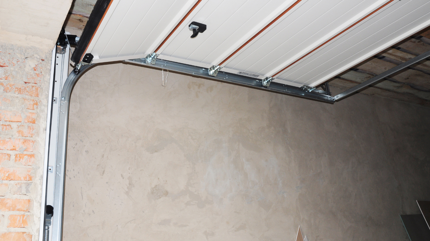 A close-up view of a white, paneled garage door mechanism showing the metal track and roller system against a plain wall.