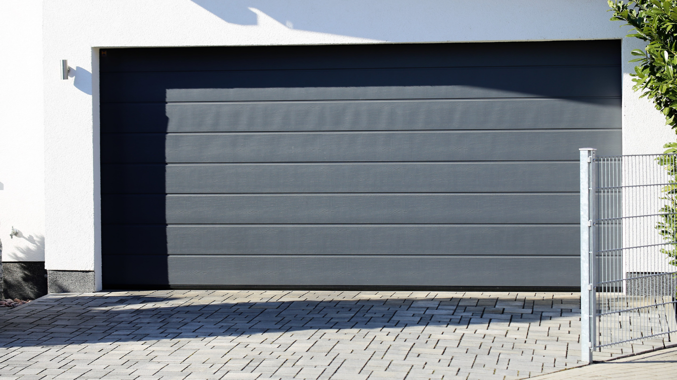 A gray, horizontal-paneled garage door installed on a modern white house with a paved driveway in front.