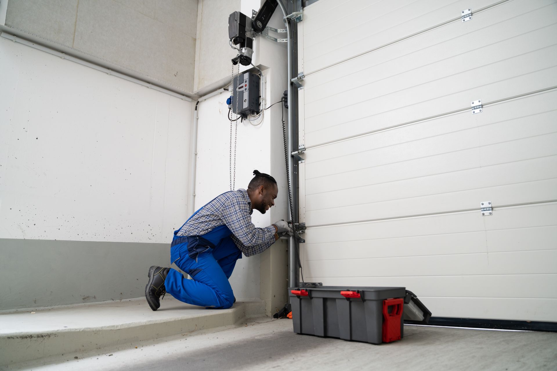 A technician in blue overalls kneeling and repairing a commercial sectional garage door with a toolbox nearby.