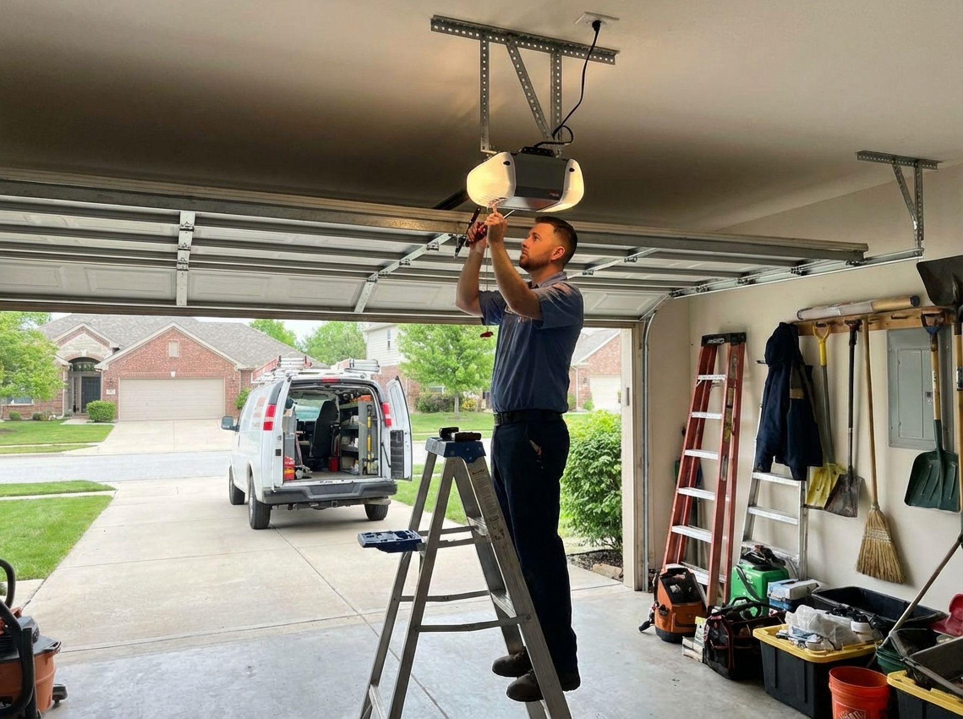 A professional technician standing on a ladder in a garage, repairing a ceiling-mounted garage door opener.