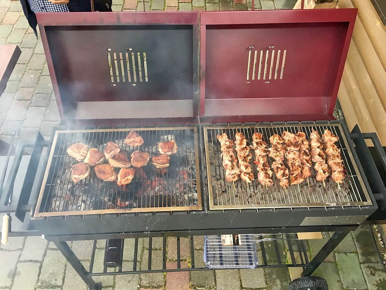 Two grills are cooking meat on a brick sidewalk.