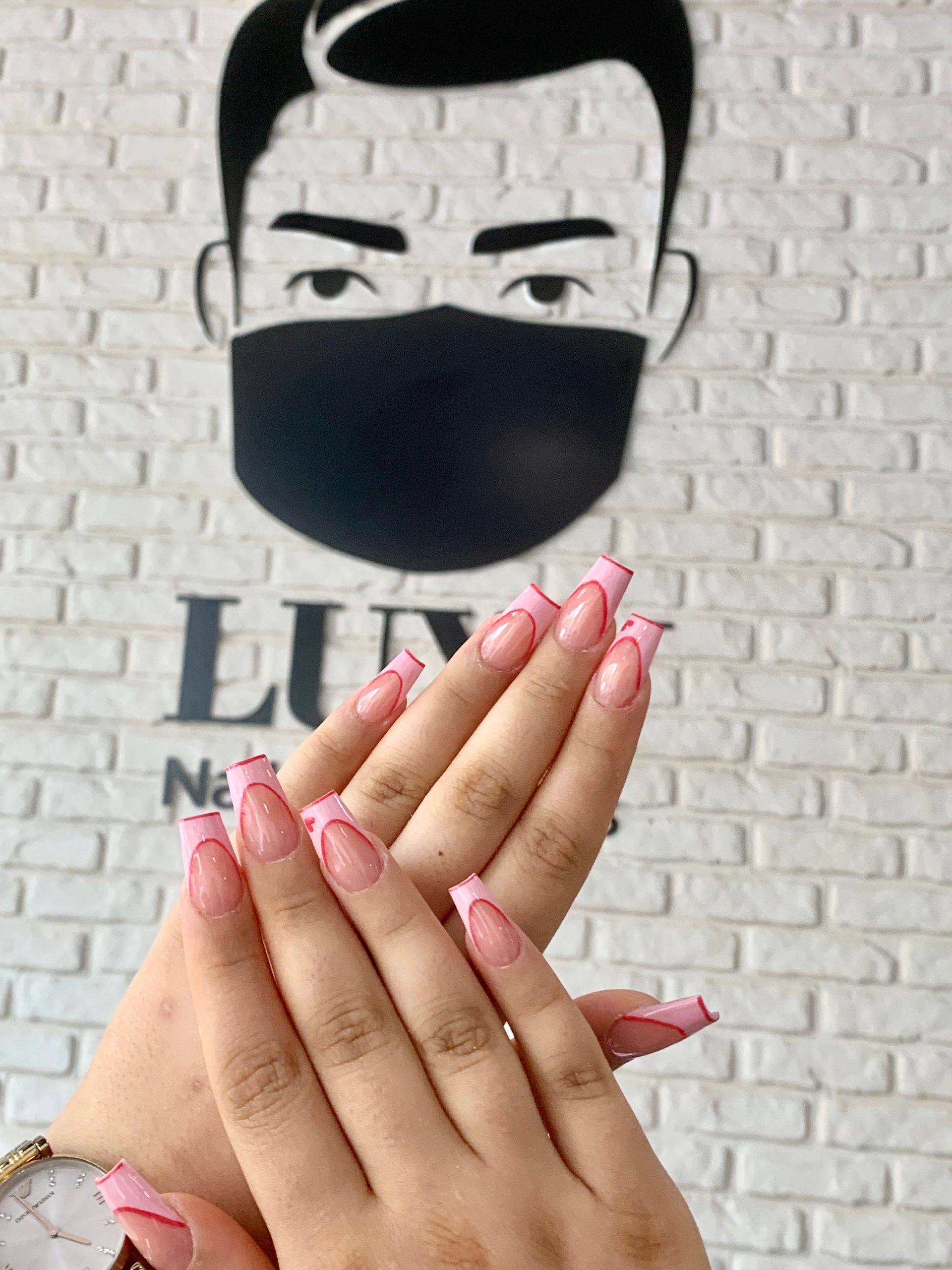 Die Hände einer Frau mit rosa Nägeln vor einer Backsteinmauer, neben der ein Mann eine Maske trägt.