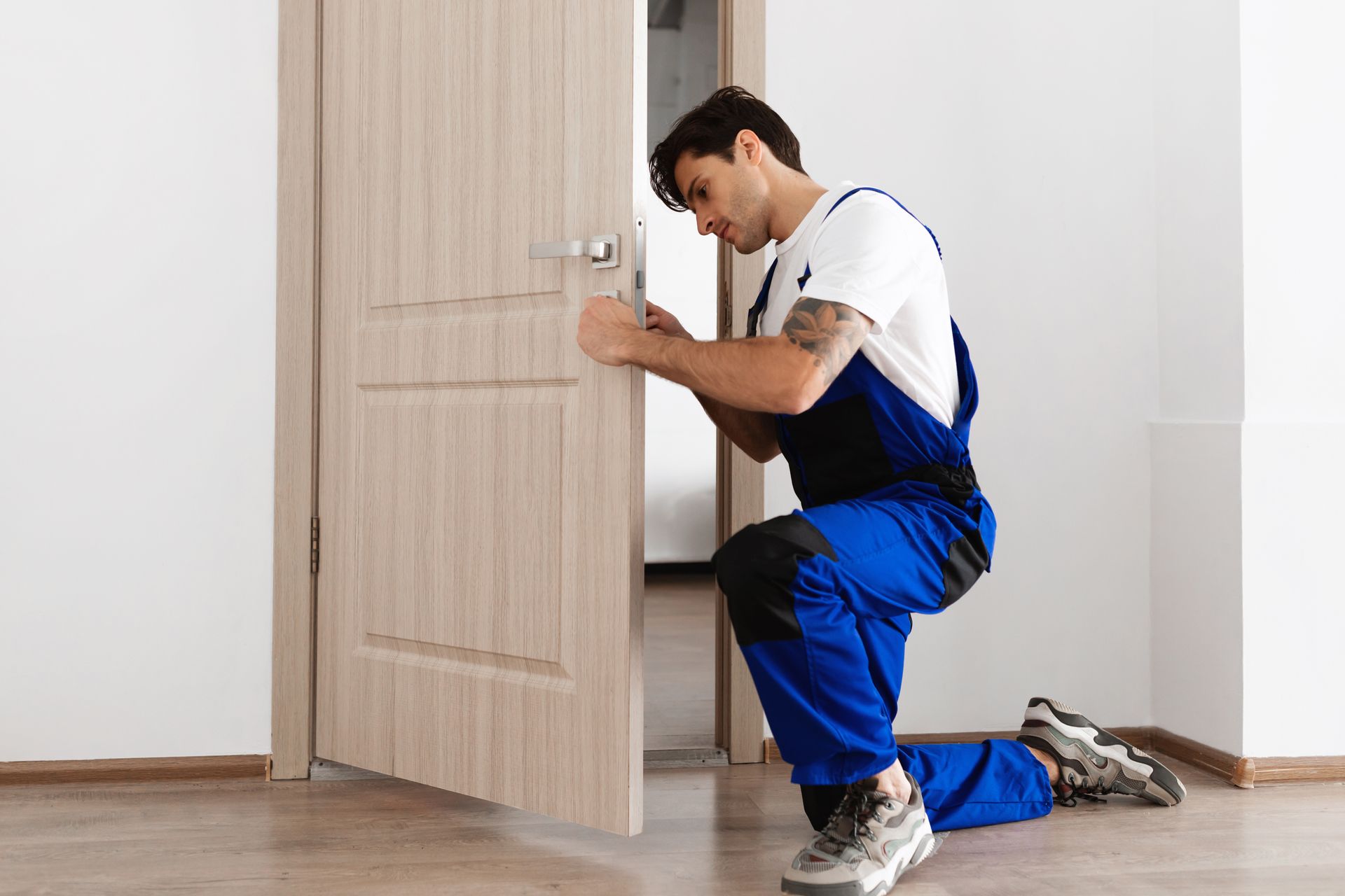 A locksmith in a blue uniform is kneeling while installing a doorknob on a wooden entrance door. A locksmith in a blue uniform is kneeling while installing a doorknob on a wooden entrance door.