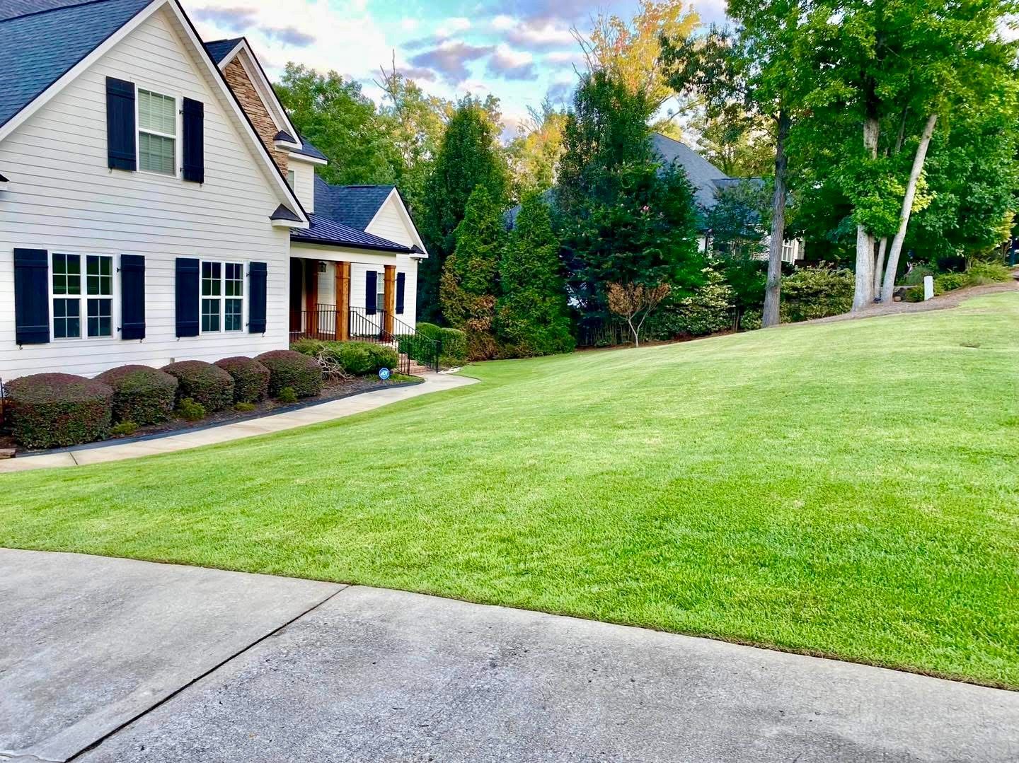A house with black shutters and a well-manicured lawn under a partly cloudy sky. Lush green grass slopes down to a concrete driveway.