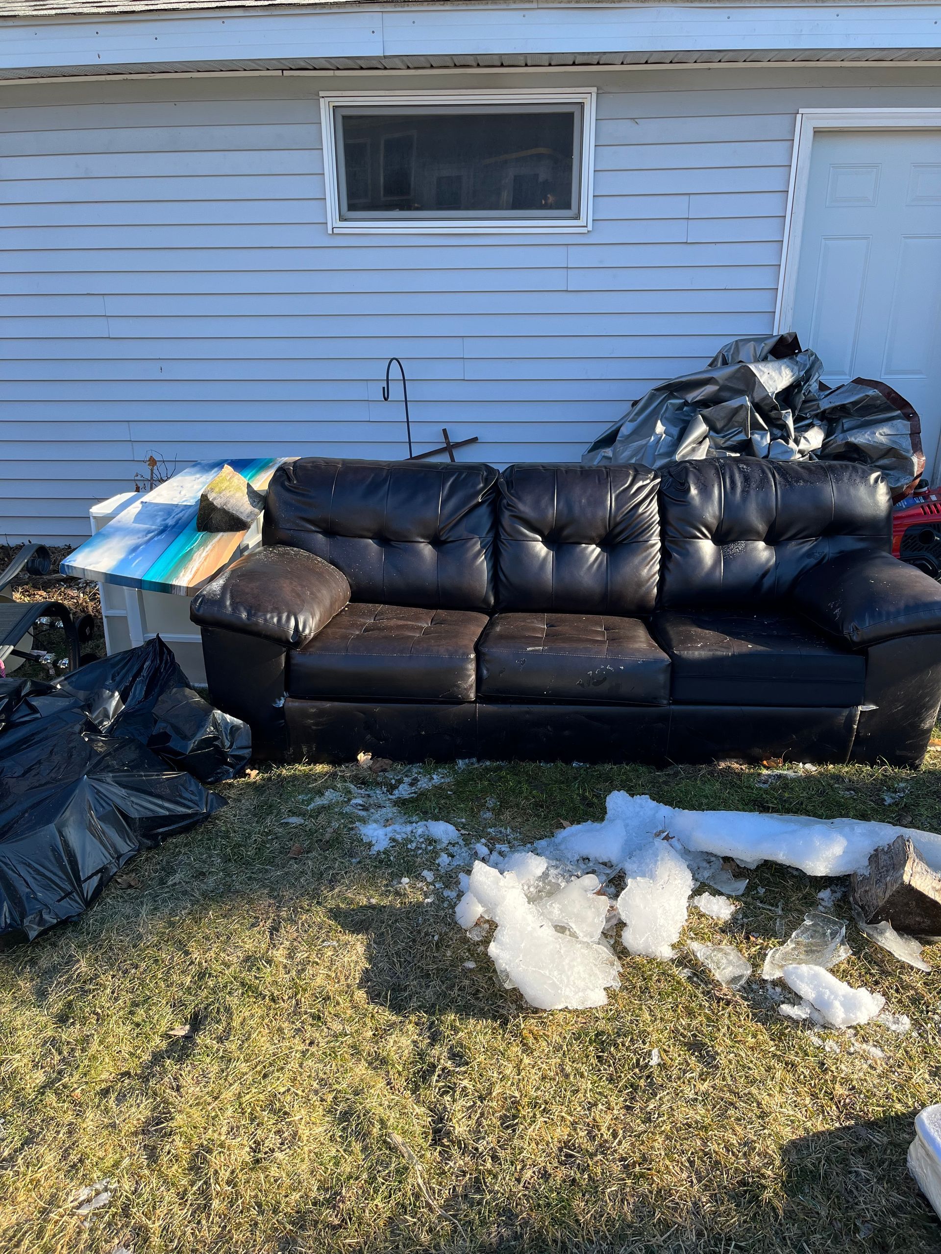 a old, ripped, black leather couch is sitting in the backyard of a house. 