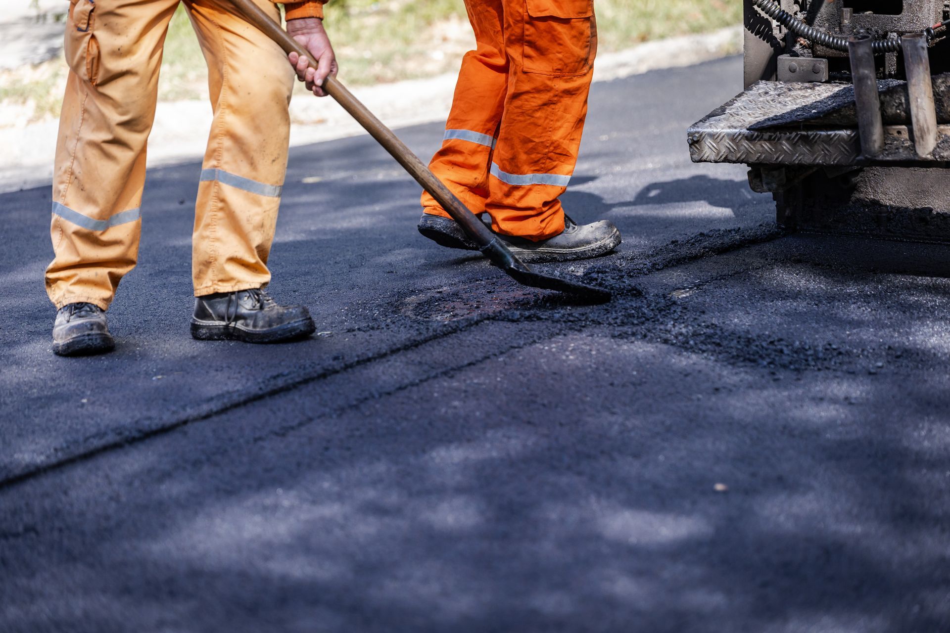 Two workers in orange pants spread asphalt with shovels on a road.