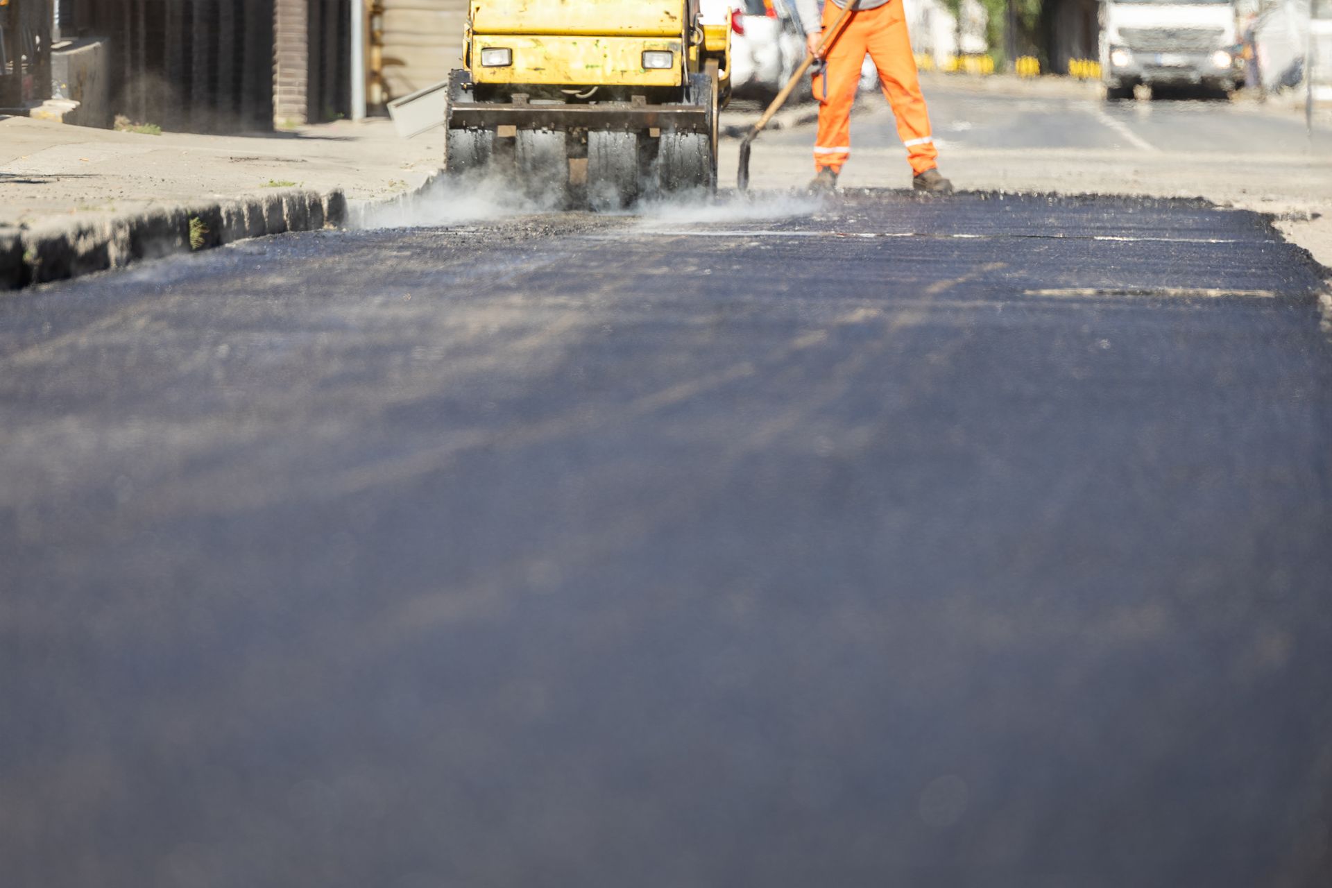 Road repair in progress; asphalt roller and worker with rake on a street, yellow and orange colors.
