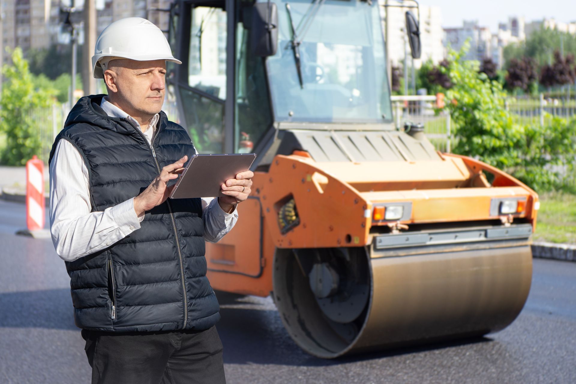 Construction worker in hard hat and vest holding a tablet, road roller in background.