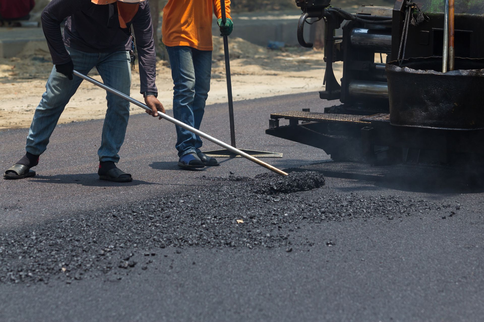 Workers paving a road, leveling asphalt with rakes near a machine.