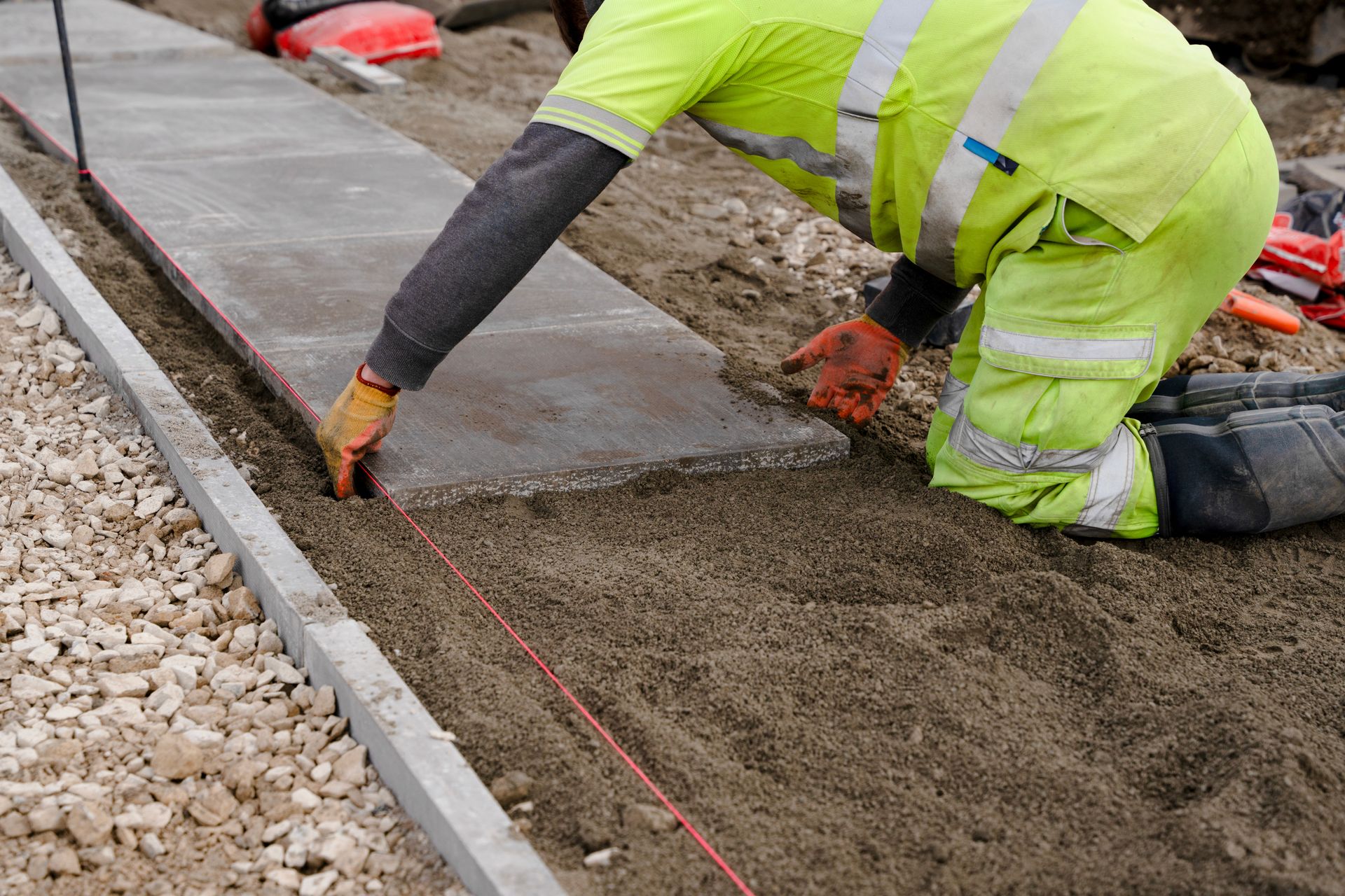 Construction worker laying a stone pavement on a sand base, guided by a string, wearing safety gear.