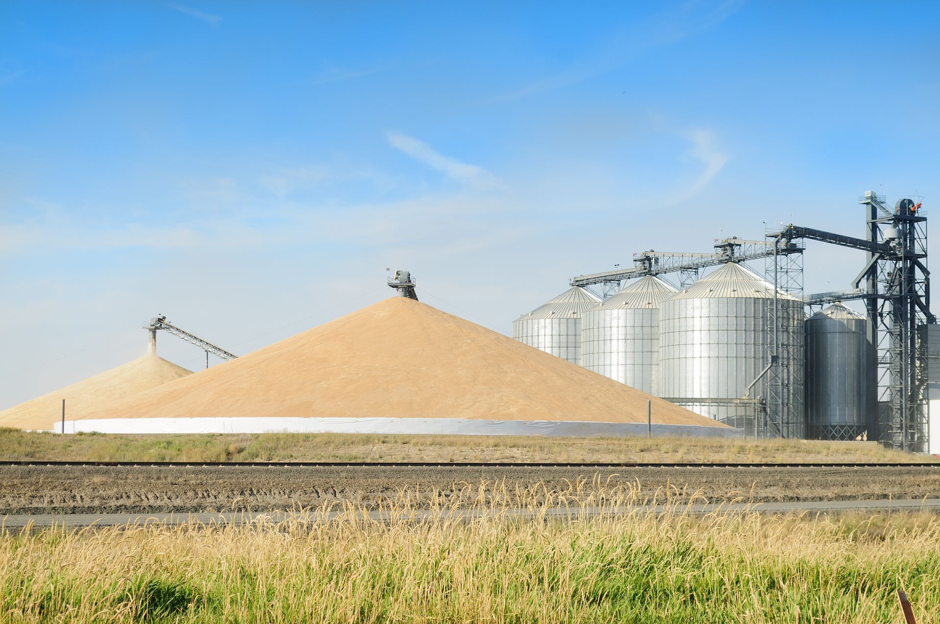 Grain silos and large pile of grain near railroad tracks under a blue sky.