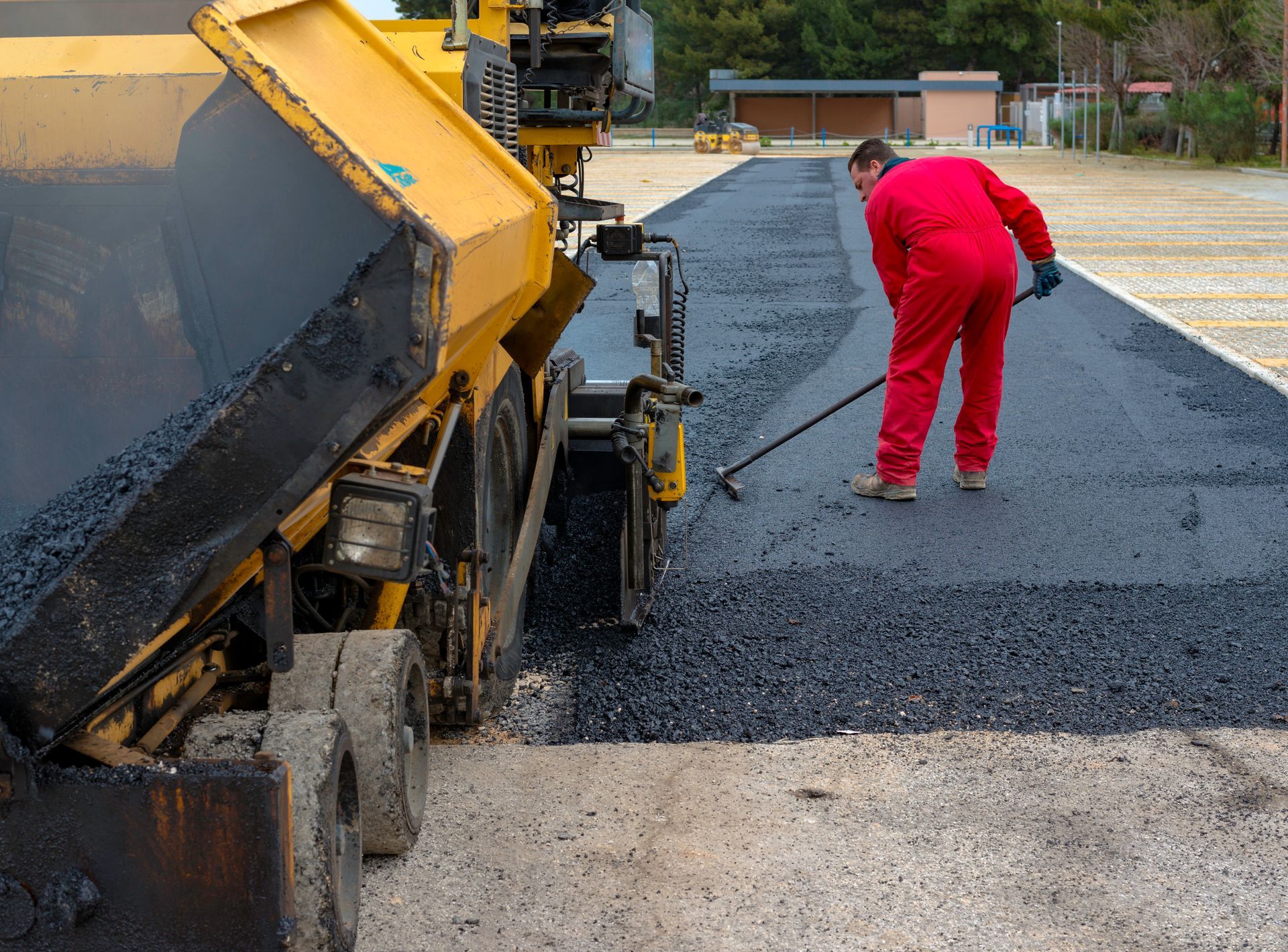 Asphalt paving machine laying asphalt; worker raking the fresh asphalt on a parking lot.