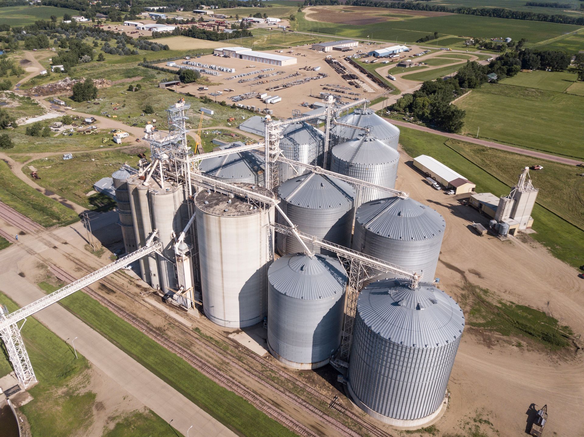 Aerial view of a grain storage facility with multiple silos, conveyor belts, and surrounding buildings on a green landscape.