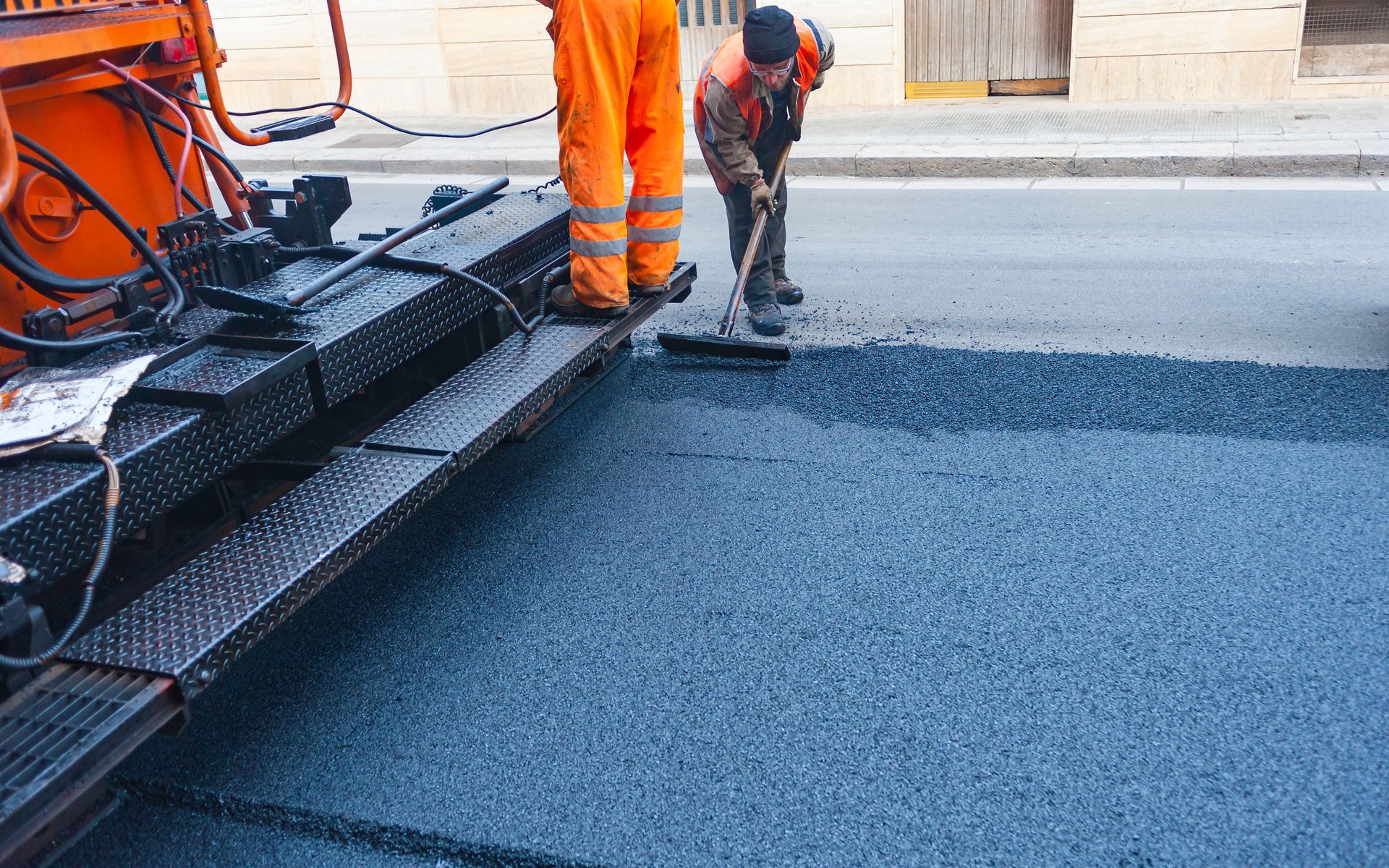 Workers paving a road with asphalt; one rakes, the other stands by the machine in orange overalls.