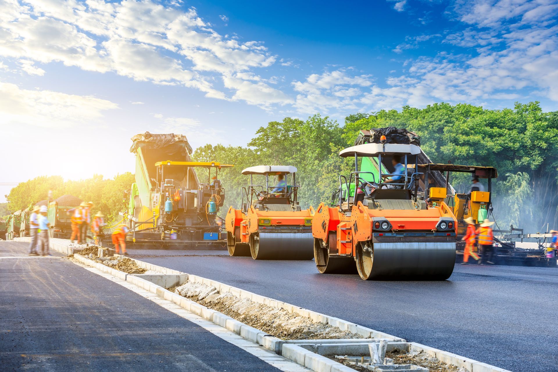 Road paving in progress, with asphalt machinery and workers in orange vests on a sunny day.