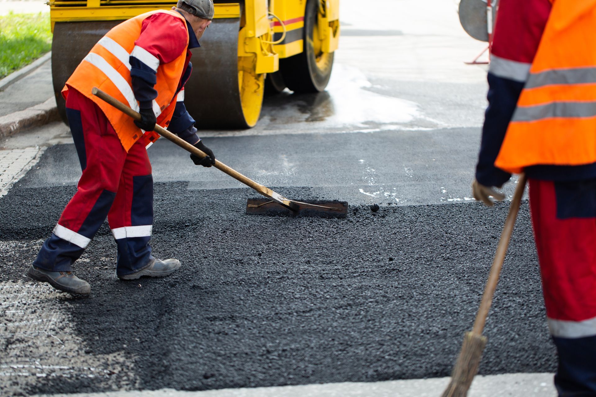 Workers in orange vests, spreading asphalt on a road, with a steamroller in the background.