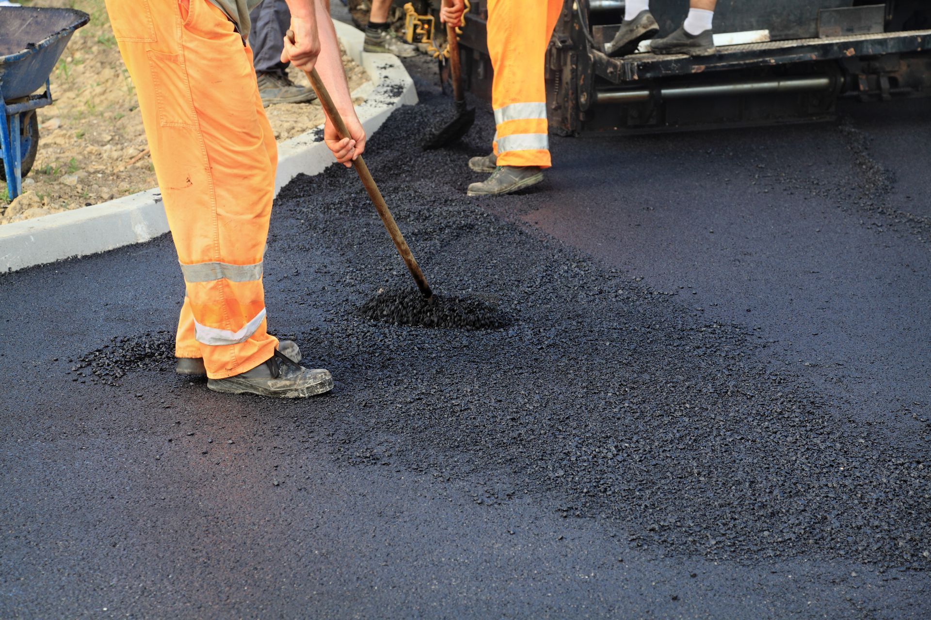 Workers in orange vests paving a road with a dark asphalt mixture using shovels and machinery.