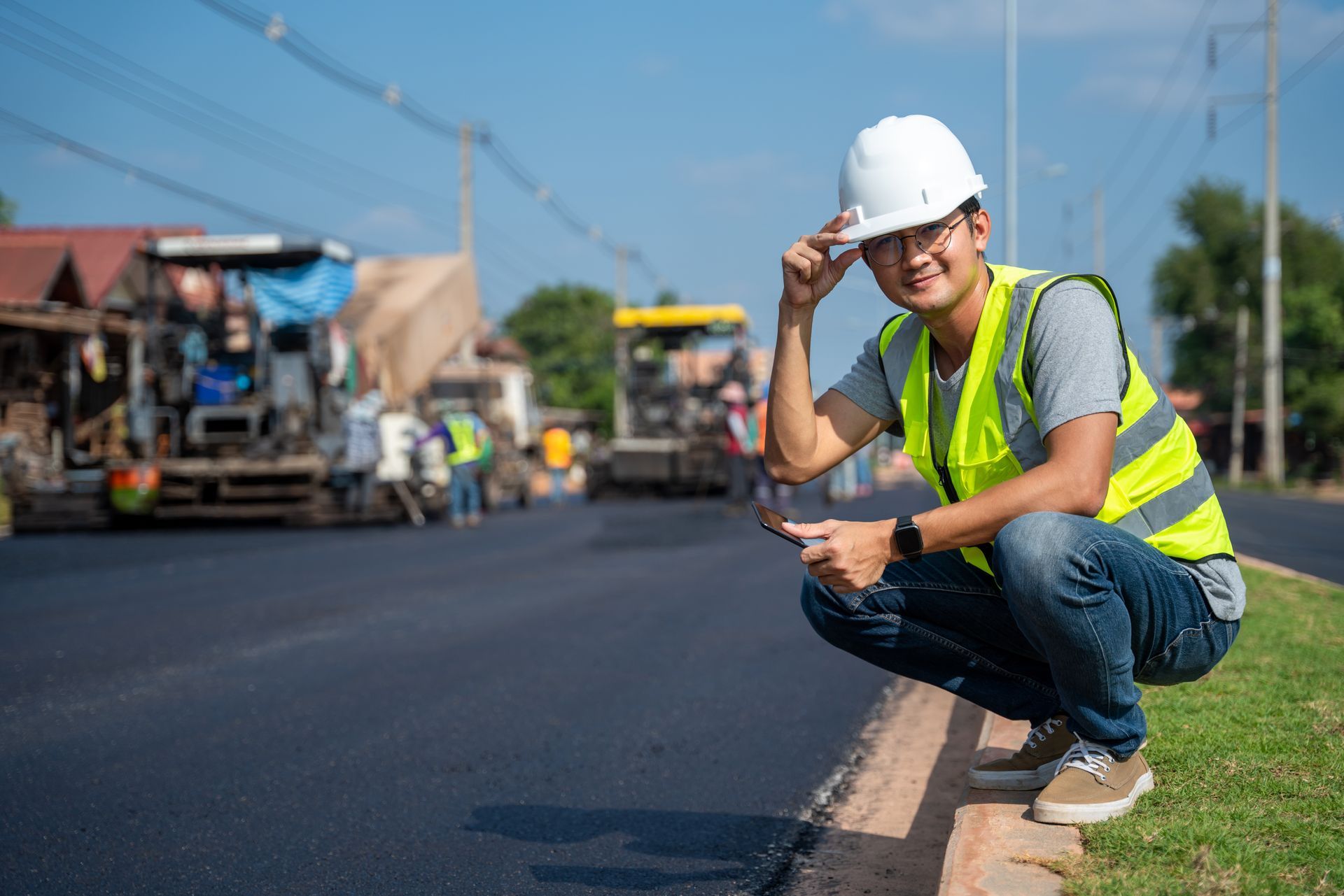 Construction worker kneels at roadside, wearing hardhat and safety vest, with paving equipment in the background.