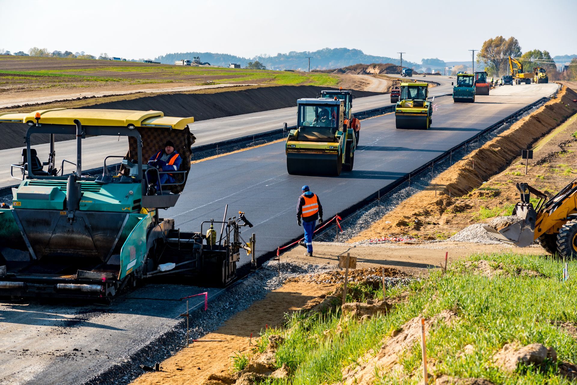 Road construction: Asphalt paver and rollers on a newly paved road, workers in orange vests, sunny day.