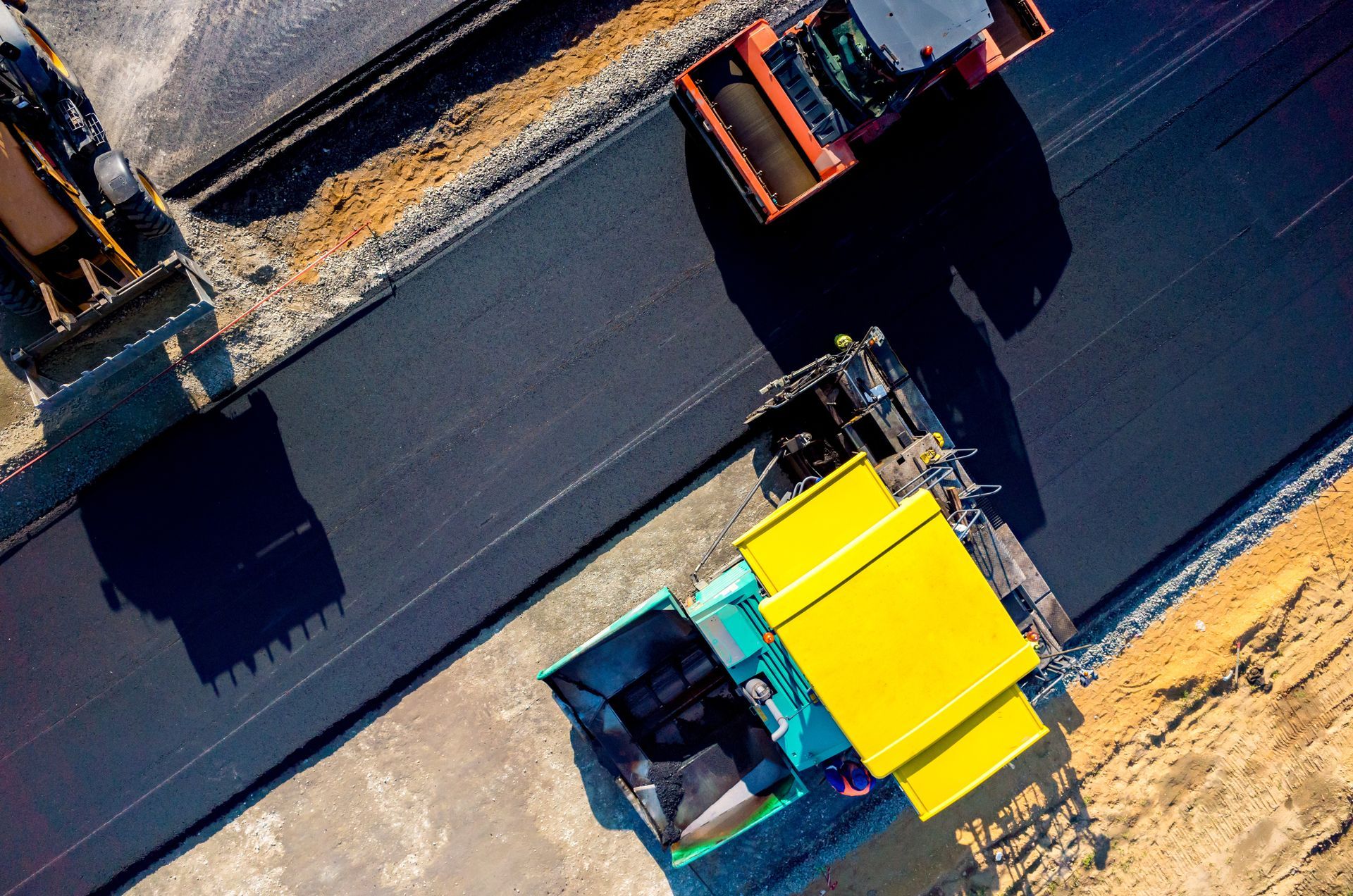 Overhead view of road construction; paving machine laying fresh asphalt, followed by a roller, on a newly built road.