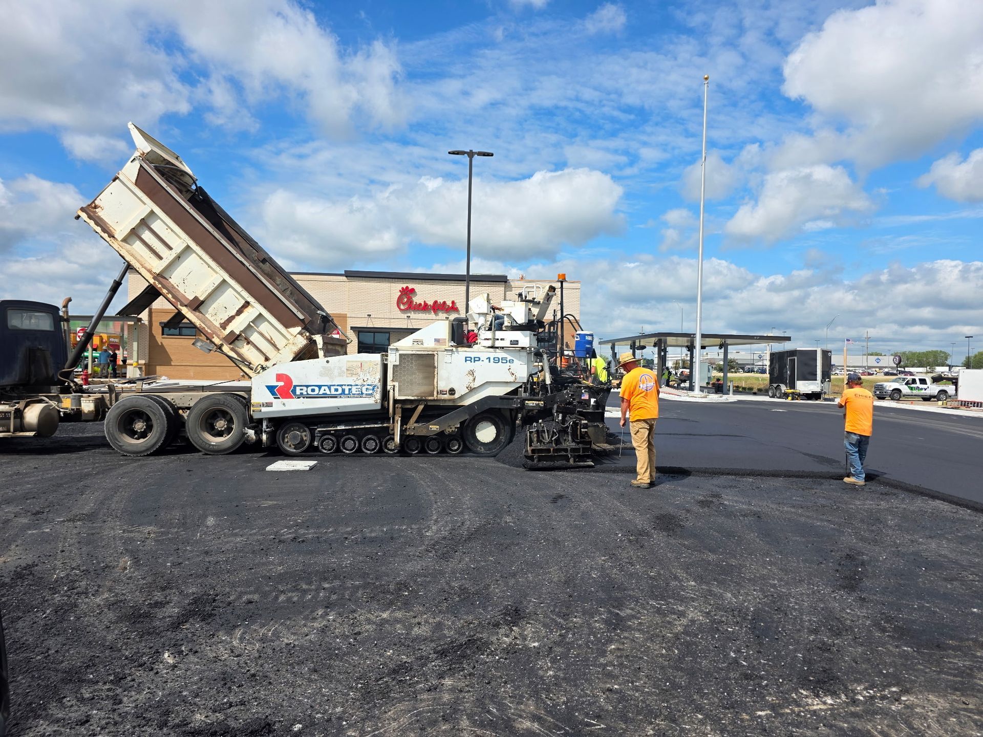 Asphalt paving at a Chick-fil-A. A dump truck unloads asphalt for a paving machine, workers watch. Blue sky.