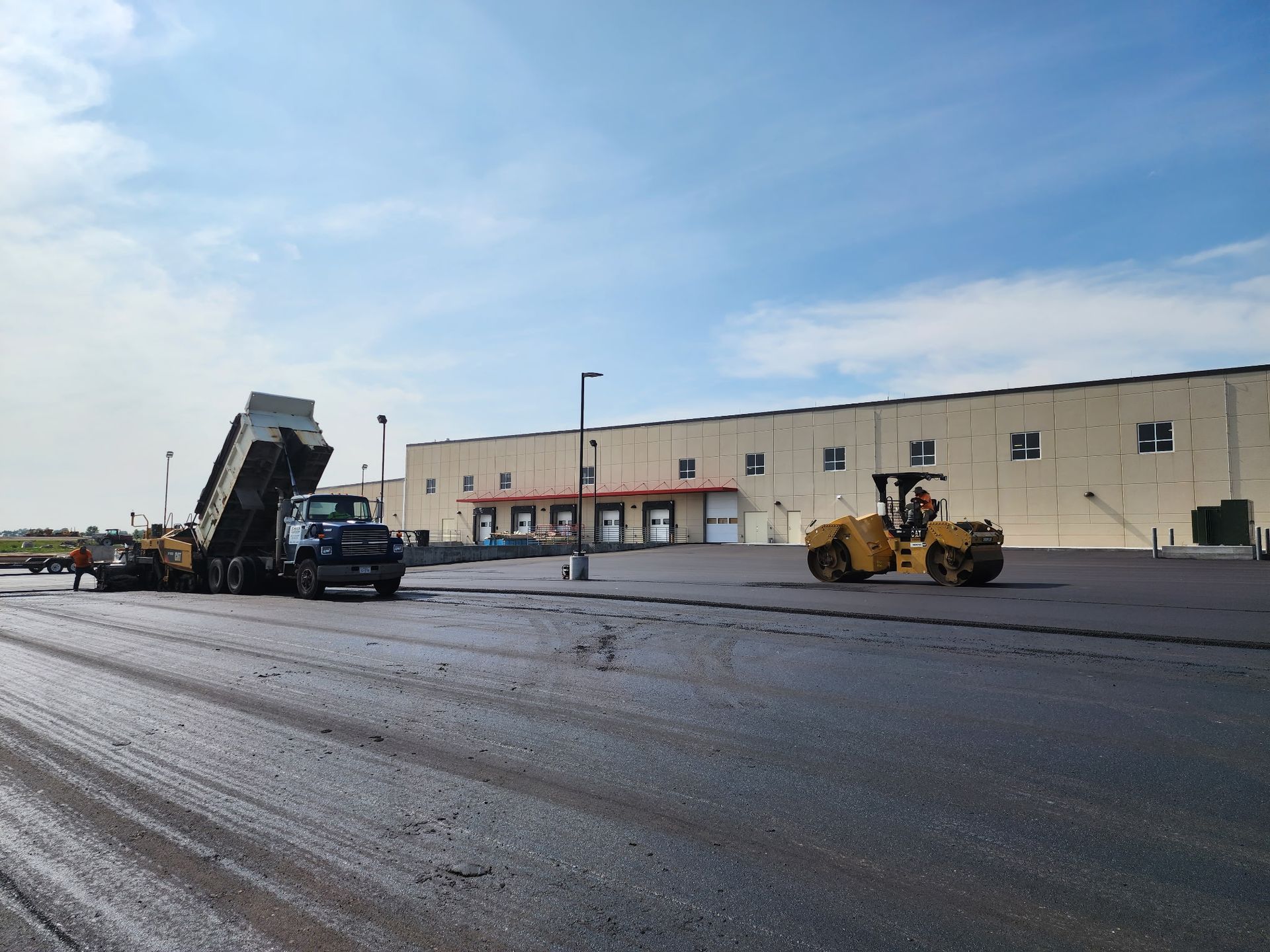 Asphalt paving operation: Dump truck unloading, yellow roller compacting, in front of a warehouse.